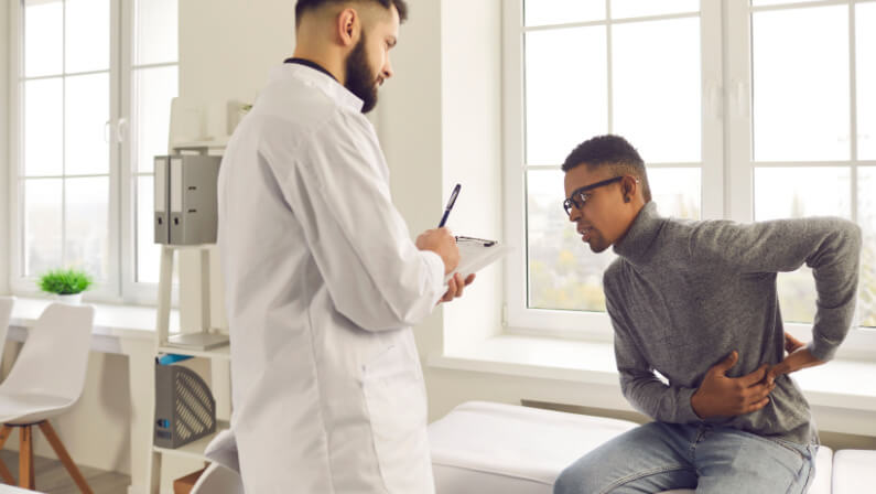 Patient telling physician about his pain and health problems during visit to hospital. Young African-American man complaining about back ache while sitting on examination bed at the doctor's office
