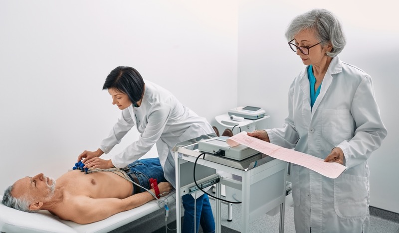 Elderly male patient receives heart rate monitored using electrocardiogram equipment with two doctors cardiologist