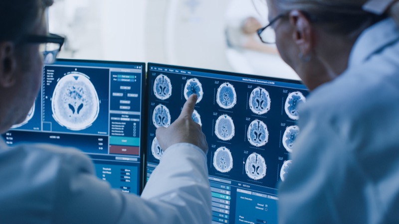 In Control Room Doctor and Radiologist Discuss Diagnosis while Watching Procedure and Monitors Showing Brain Scans Results, In the Background Patient Undergoes MRI or CT Scan Procedure.