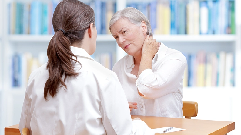 Senior female patient consulting her physician or doctor on account of chronic neck pain caused by fibromyalgia, osteoporosis or a herniated disc.