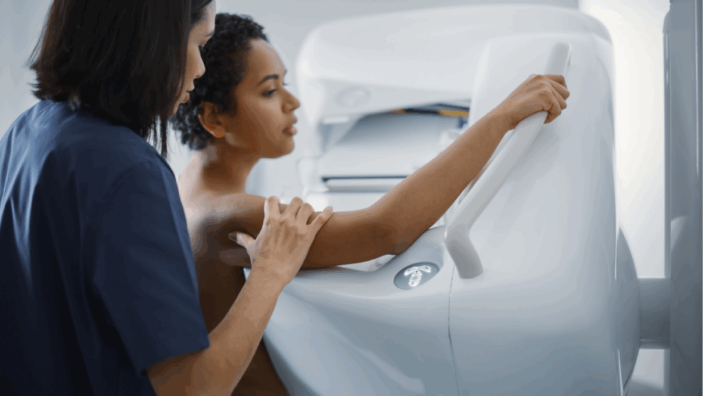 Friendly Female Doctor Explains the Mammogram Procedure to a Topless Latin Female Patient with Curly Hair Undergoing Mammography Scan. Healthy Female Does Cancer Prevention Routine in Hospital Room.