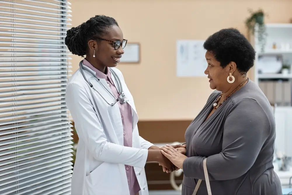 Side view of cheerful Black woman doctor greeting mature female patient while shaking hands in clinic